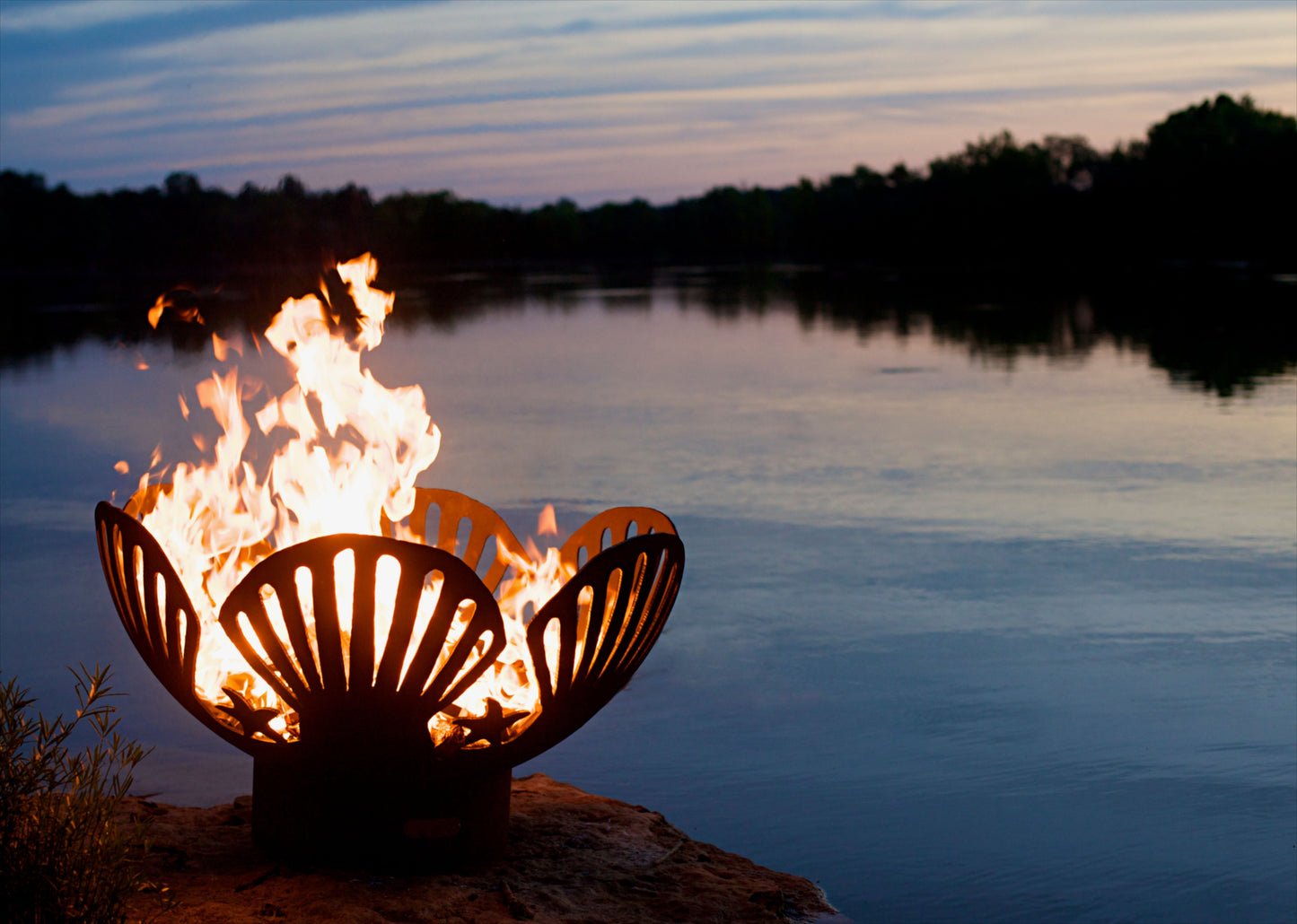 The beautiful Barefoot Beach Fire Pit, displayed on the lake shore, with a warm roaring flame.