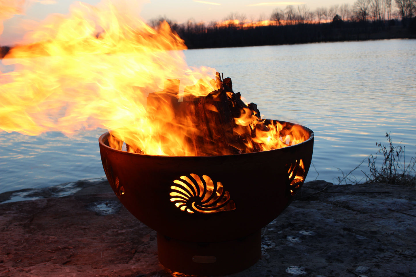 The BeachComber Fire Pit, displayed on the lake shore, burning firewood with a huge flame.