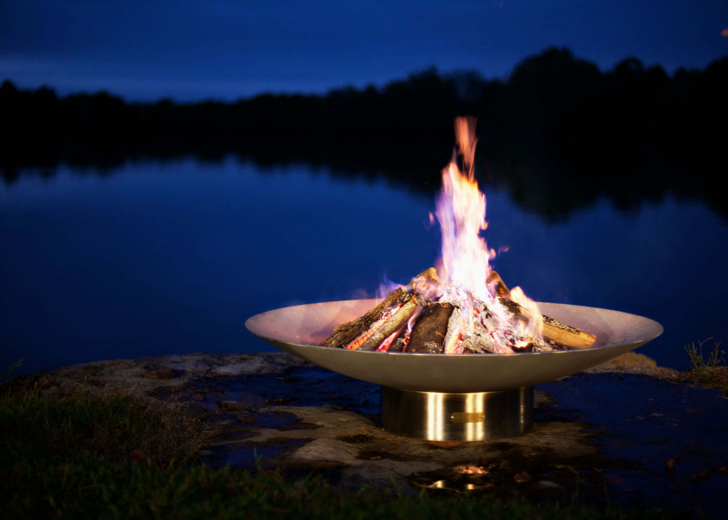 The Bella Vita Series Fire Pit, Displayed lakeside, with a huge burning flame at night.