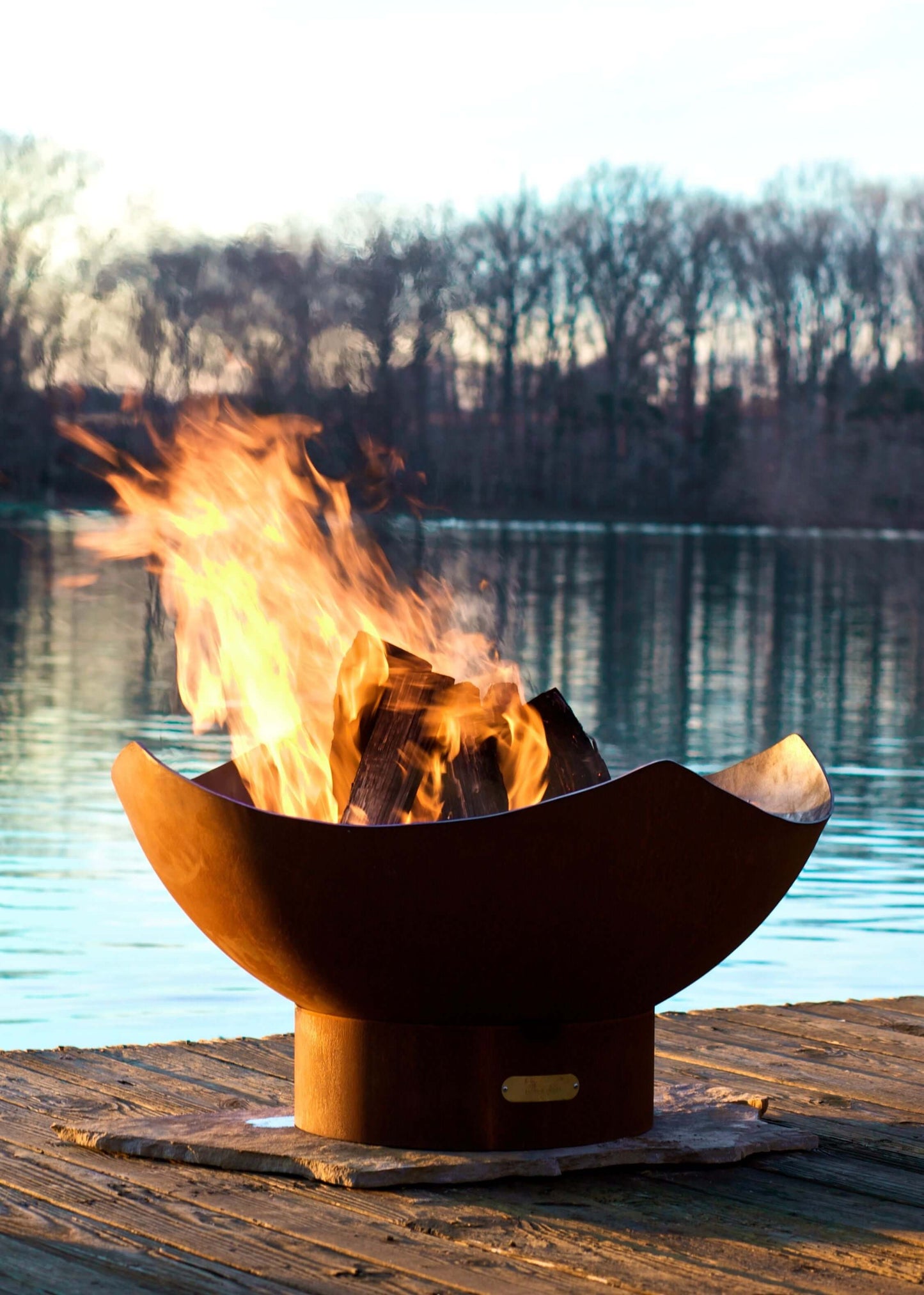 The elegant Manta Ray Fire Pit displayed on a lakeside dock, burning wood with a warm flame.
