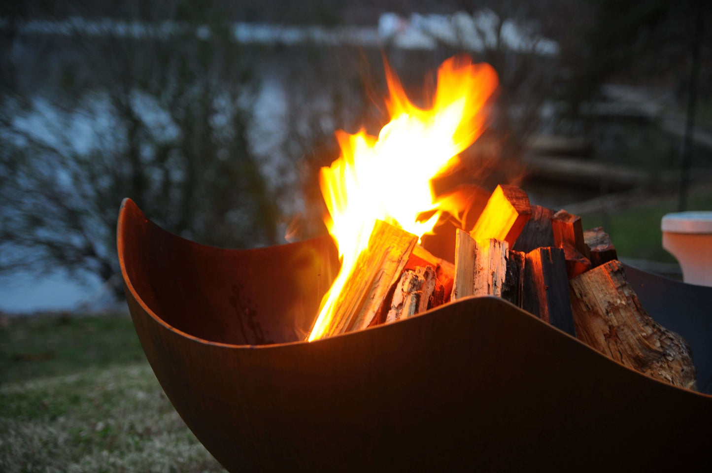 The elegant Manta Ray Fire Pit displayed with a warm fiery flame, on the lakeside grass.