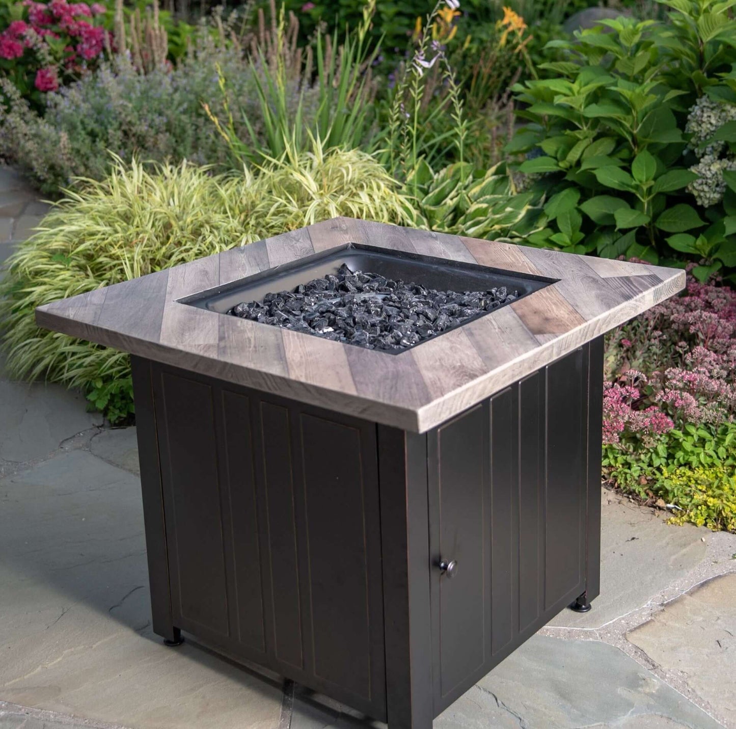 The Harper, 30" Square Gas Outdoor Fire Pit with Printed Cement Resin Mantel, displayed in a backyard garden.