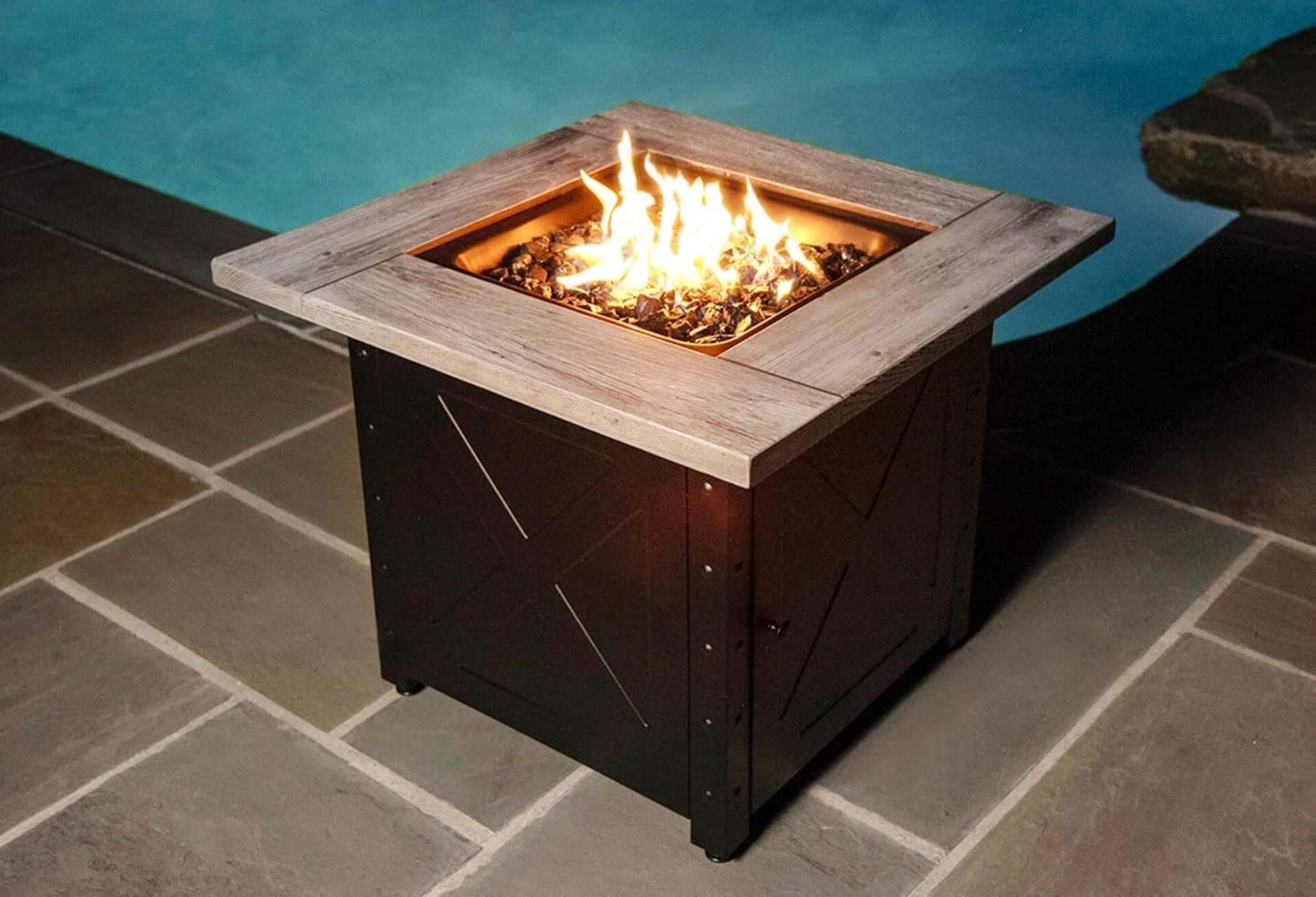 The Mason, 30" Square Gas Outdoor Fire Pit with Printed Wood Lat look Cement Resin Mantel, displayed on a pool side patio, with a warm flame.