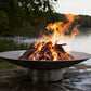 The Bella Vita Series Fire Pit, Displayed lakeside, with a huge burning flame.