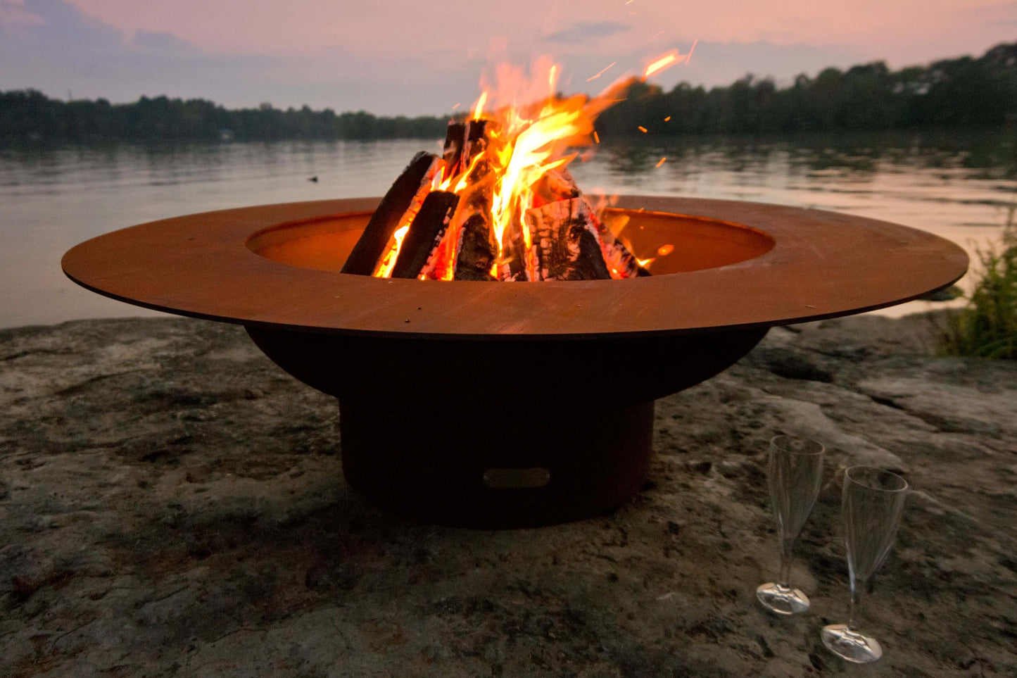 The stunning Magnum Fire Pit, Displayed on a lakeside rock, with a warm roaring flame.
