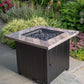 The Harper, 30" Square Gas Outdoor Fire Pit with Printed Cement Resin Mantel, displayed in a backyard garden.